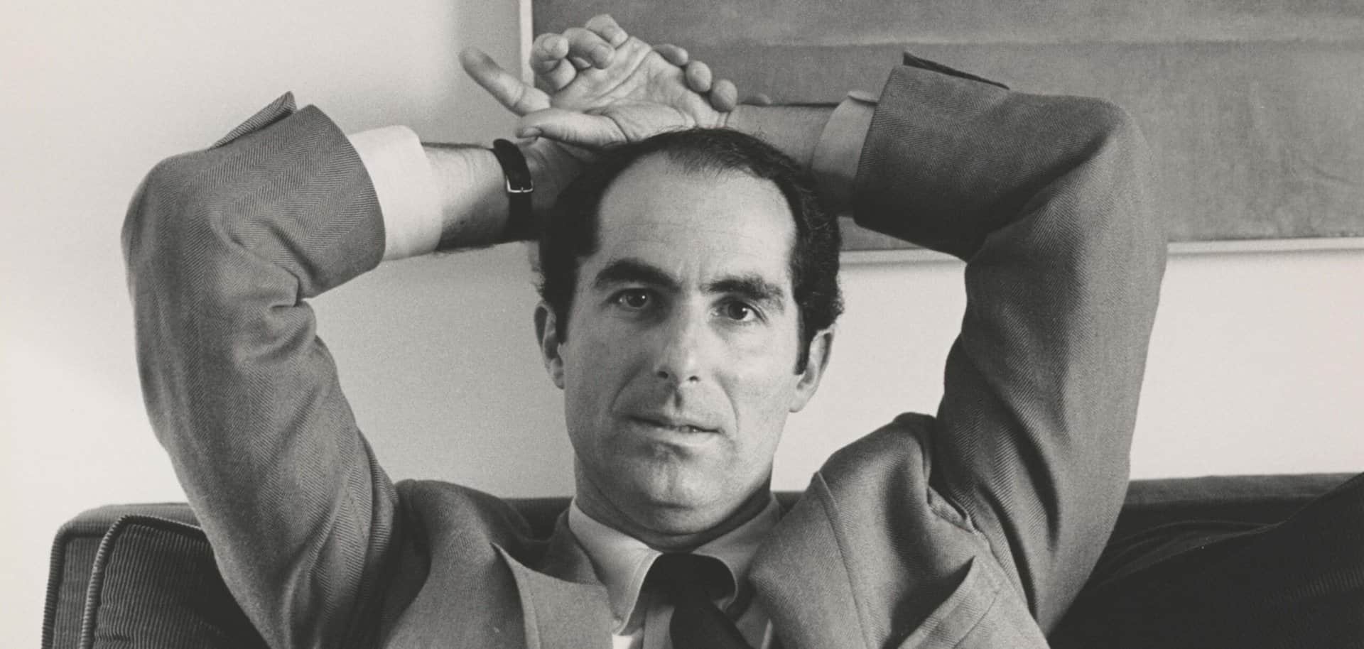 A greyscale close-up photo of Philip Roth staring at the camera, wearing a suit.