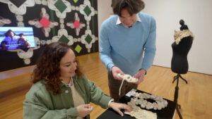 Jason Strother and Amanda Cachia stand together in the exhibition space for Smoke and Mirrors at the Zimmerli Art Museum. They are at a table touching textile artworks with their hands and smiling.