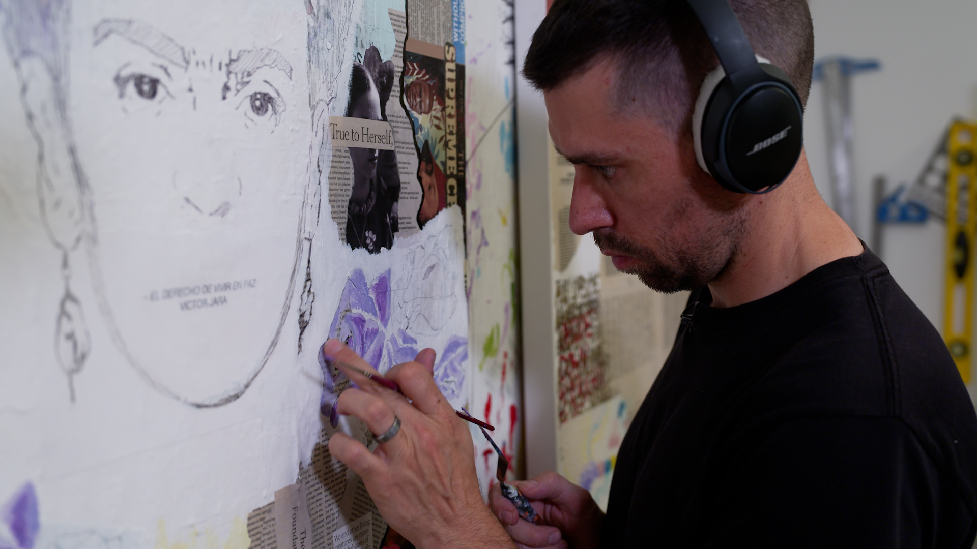 An up-close view of Lawrence Ciarallo painting in his studio. He wears headphones and is working on a collage. He does not look at the camera and is focused on his artwork.